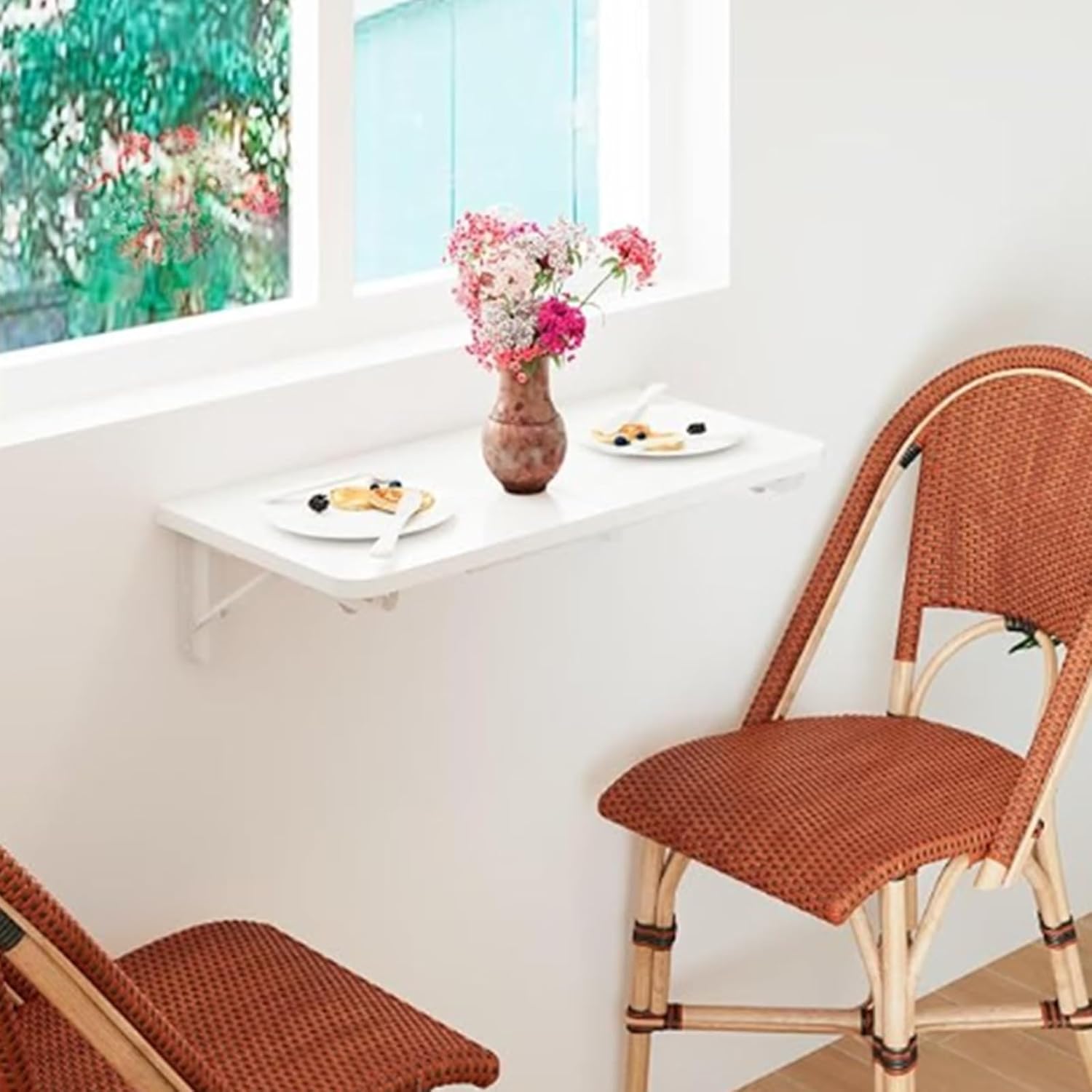 A wall-mounted drop-leaf table folded down, holding a laptop and a teacup, on a concrete balcony. A foldable stool and a hanging planter add charm, with an urban view.