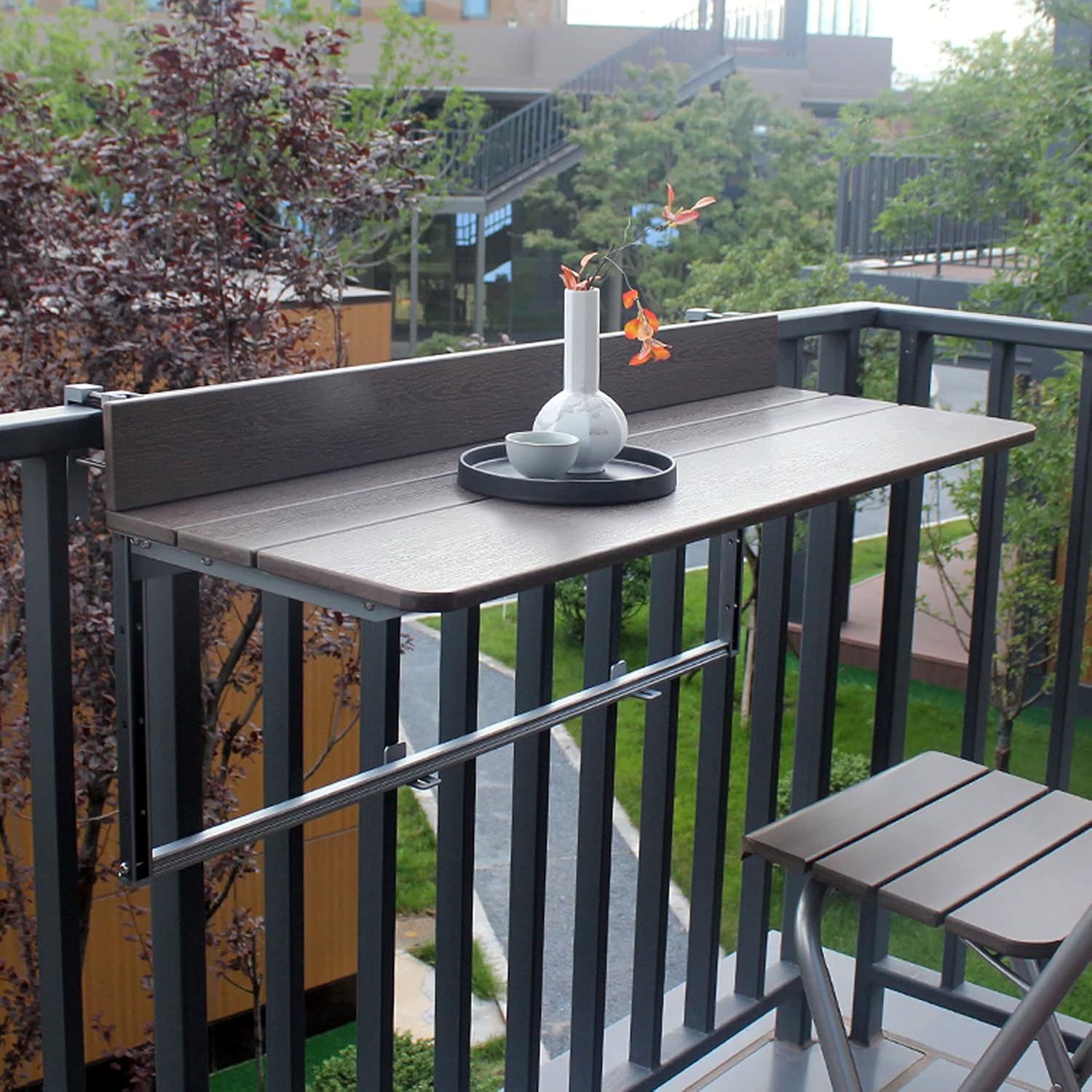 A wall-mounted bar counter with two foldable bar stools, holding wine glasses, on a concrete balcony. A hanging herb planter and a city view create a chic urban scene.