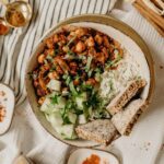A bowl of creamy cashew pasta with vibrant spinach, twirled on a fork, on a white tablecloth. A jar of nutritional yeast and a lemon wedge sit on a rustic wooden table, with a cozy kitchen backdrop.