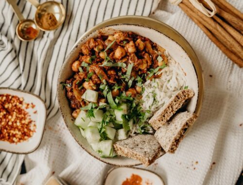A bowl of creamy cashew pasta with vibrant spinach, twirled on a fork, on a white tablecloth. A jar of nutritional yeast and a lemon wedge sit on a rustic wooden table, with a cozy kitchen backdrop.